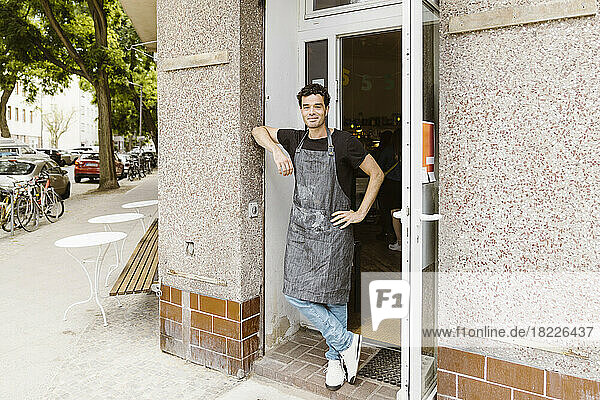 Lächelnder männlicher Café-Besitzer mit der Hand auf der Hüfte  der sich an die Wand am Eingang lehnt