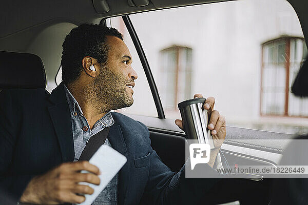 Lächelnder männlicher Pendler mit isoliertem Getränkebehälter und Smartphone  der aus dem Autofenster schaut
