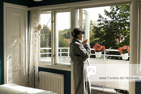 Side view woman wearing bathrobe having tea while leaning on balcony doorway in bedroom