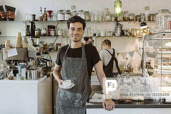 Lächelnder männlicher Besitzer mit Schürze  der eine Kaffeetasse in einem Café hält