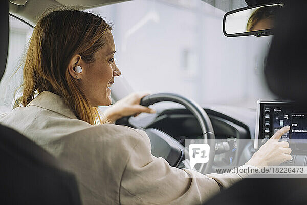 Lächelnde Geschäftsfrau mit drahtlosen In-Ear-Kopfhörern  die das Armaturenbrett im Auto bedient