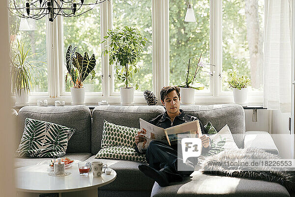Junger Mann liest Zeitung  während er zu Hause auf dem Sofa im Wohnzimmer sitzt