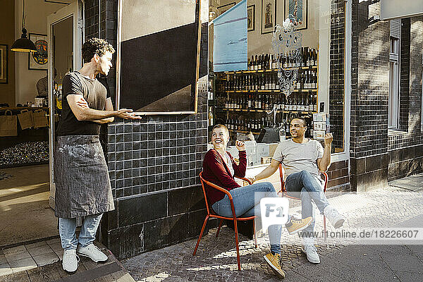 Männlicher Besitzer im Gespräch mit Kunden  die auf einem Stuhl auf dem Gehweg vor dem Geschäft sitzen