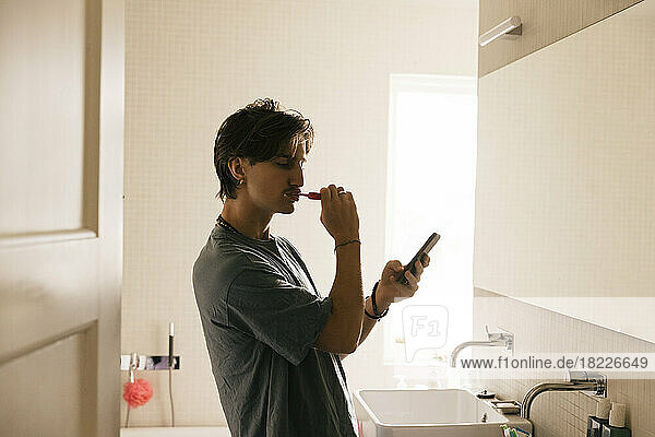 Seitenansicht eines jungen Mannes  der beim Zähneputzen im heimischen Badezimmer sein Smartphone benutzt