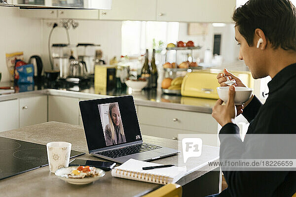 Junger männlicher Freiberufler frühstückt  während er mit einer Kollegin am Laptop in der Küche telefoniert