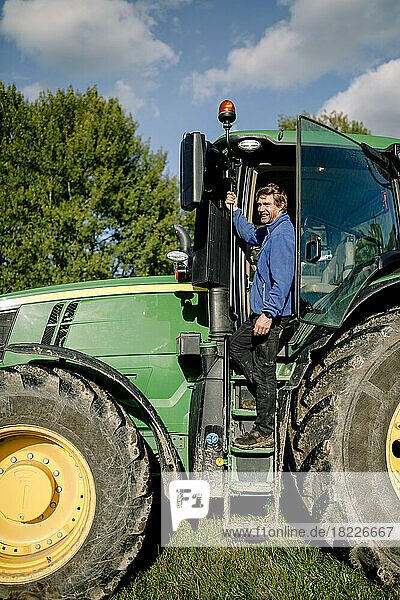 Glücklicher reifer Landwirt steht auf einem Traktor auf einem Bauernhof unter bewölktem Himmel