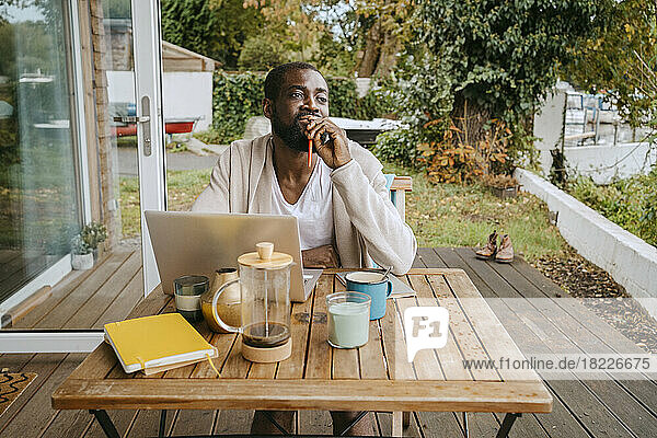 Nachdenklicher Mann mit Laptop sitzt am Tisch auf der Veranda