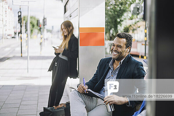 Glücklicher männlicher Freiberufler mit kabellosen In-Ear-Kopfhörern  der mit einem Tagebuch gegen eine Geschäftsfrau an der Bushaltestelle sitzt