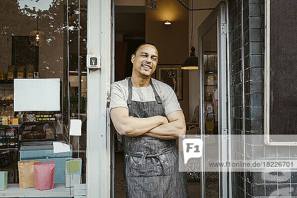 Smiling male owner leaning with arms crossed on deli doorway