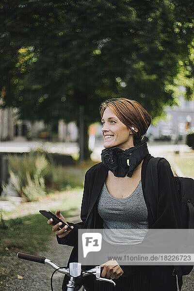 Glückliche Geschäftsfrau mit Mobiltelefon und Fahrrad beim Spaziergang im Park