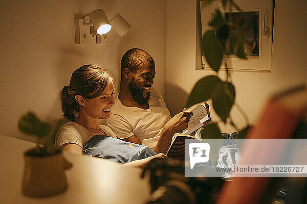 Smiling couple reading books by illuminated sconce mounted on wall at home