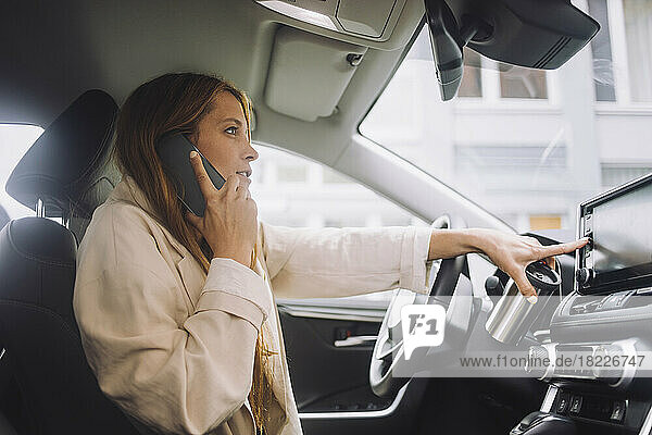 Weibliche Unternehmerin  die über ihr Mobiltelefon spricht  während sie das Touchscreen-Bedienfeld auf dem Armaturenbrett im Auto benutzt