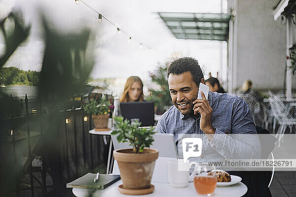 Smiling male entrepreneur talking on smart phone while sitting with laptop at cafe