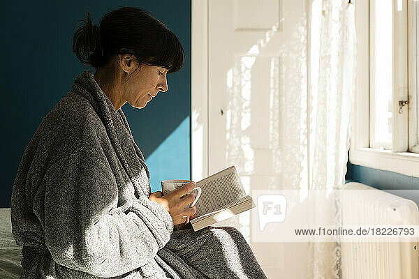 Frau im Bademantel  die eine Tasse hält  während sie im Schlafzimmer zu Hause ein Buch liest