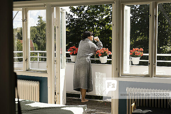 Rear view of woman enjoying tea while leaning on railing in balcony seen through doorway
