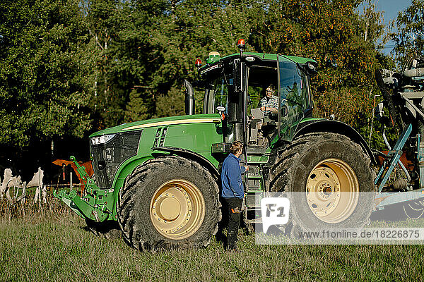 Landwirt sitzt im Traktor und diskutiert mit einem Mann  der an einem sonnigen Tag im Feld steht