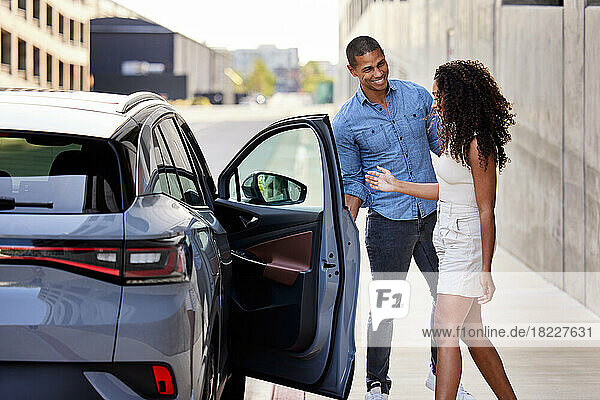 Smiling man opening door of electric car for girlfriend at roadside