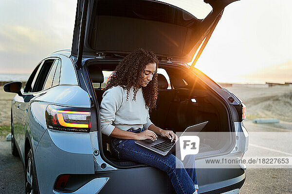 Young woman using laptop sitting in car trunk during sunset