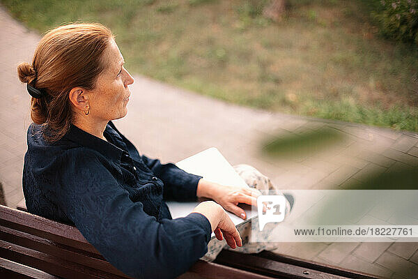 Mature business woman holding laptop sitting on bench in city