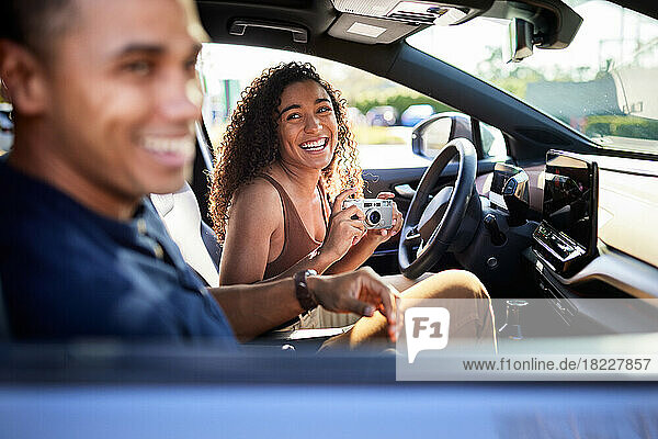 Happy woman with camera looking at boyfriend in a car during road trip