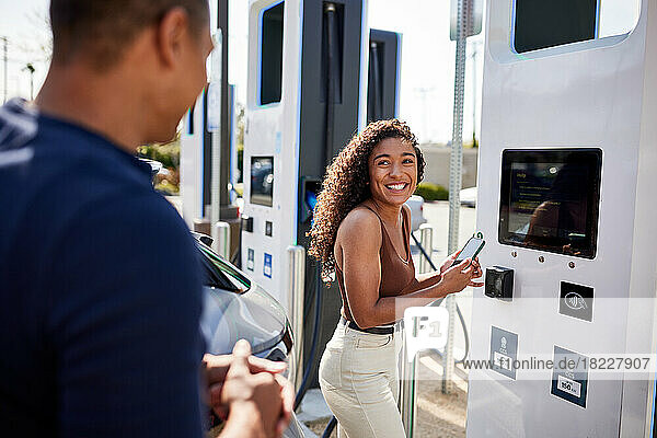 Happy woman holding smart phone talking with man at charging station