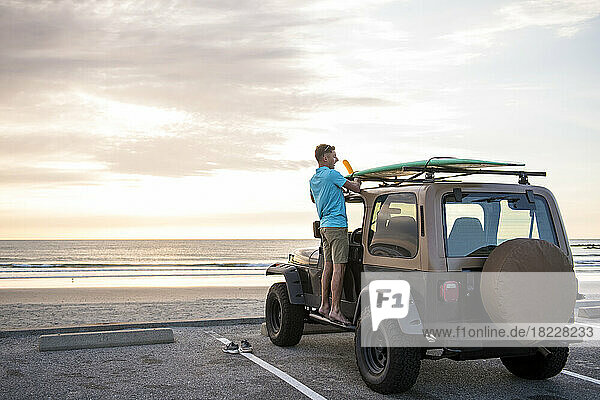 Young Man with surfboard on top of car at sunrise at Beach