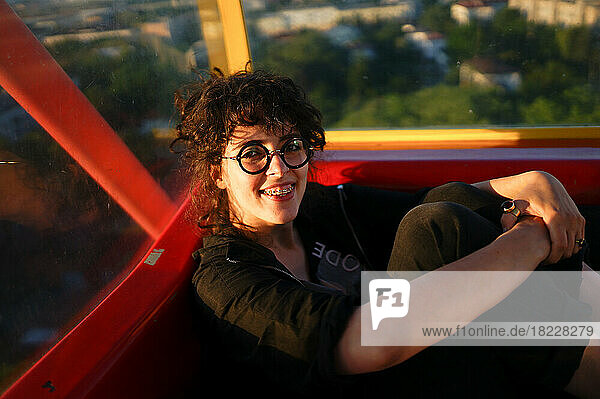 Smiling curly woman in glasses sitting in ferris wheel cabin