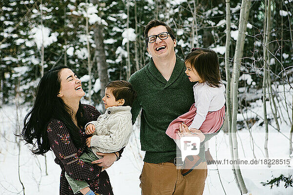 Family laughs with two small toddlers in snow forest