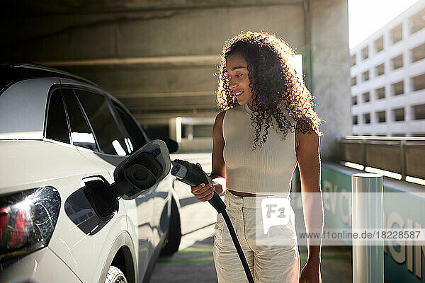 Young woman holding electric plug by car at charging station