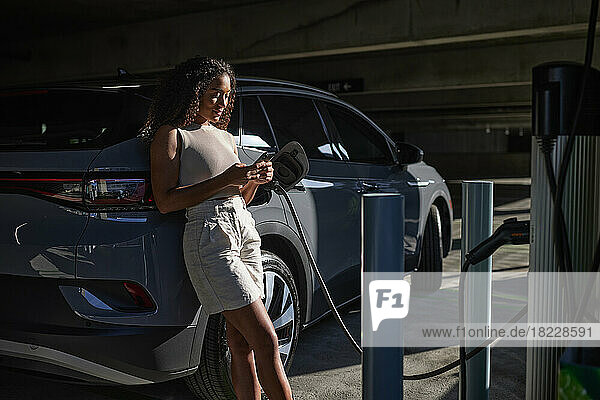 Woman text messaging leaning on electric car in parking garage