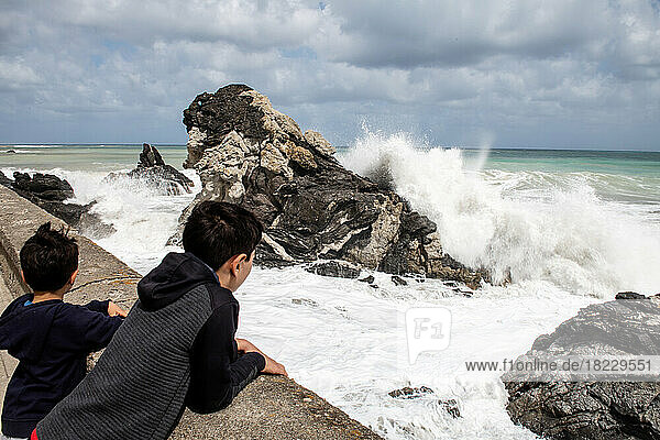 Boys (6-7  12-13) looking at sea waves crashing against rocks  Sicily  Italy