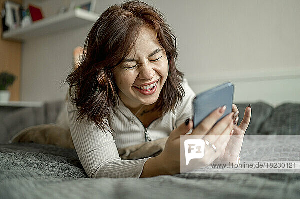 Cheerful young woman with smart phone lying on bed at home