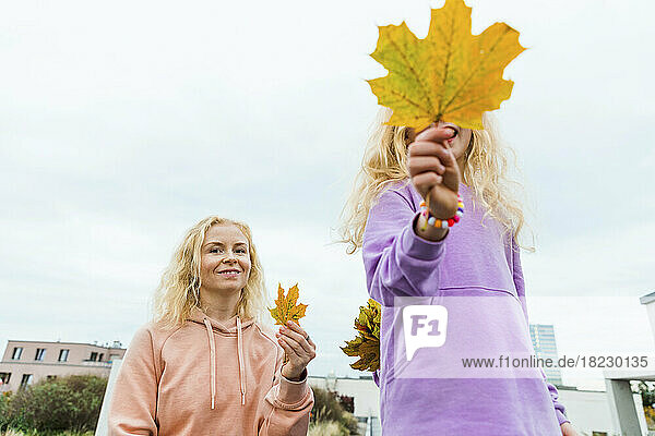 Smiling woman holding autumn leaf by daughter in front of sky