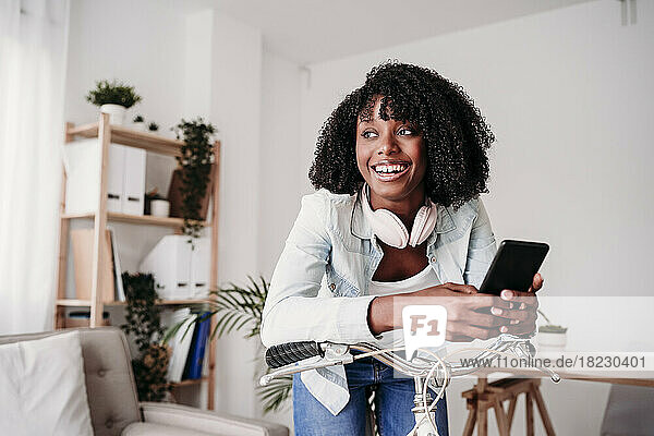 Happy woman with smart phone leaning on bicycle at home