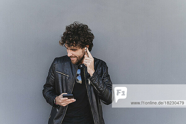 Mann hört Musik über kabellose In-Ear-Kopfhörer und hält sein Mobiltelefon an eine graue Wand gelehnt