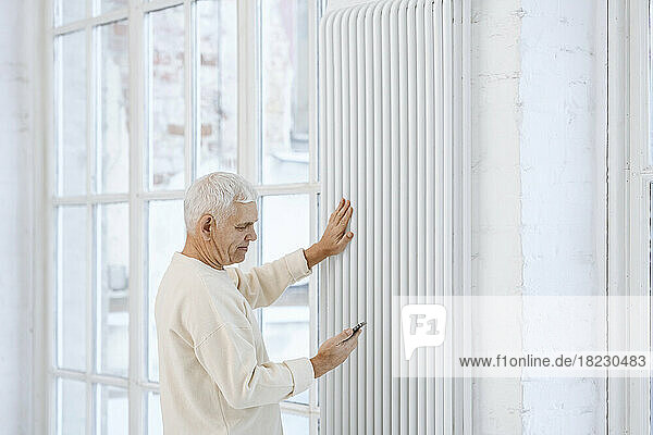 Senior man using smart phone touching radiator at home