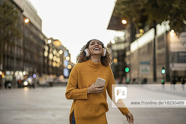 Glückliche junge Frau hält ihr Mobiltelefon in der Hand und genießt Musik  die sie über drahtlose Kopfhörer auf dem Fußweg hört