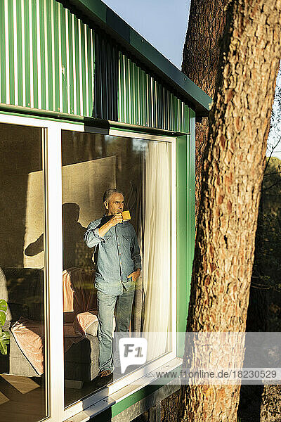 Reifer Mann steht mit einer Tasse Kaffee am Fenster eines Hauses und schaut hinaus