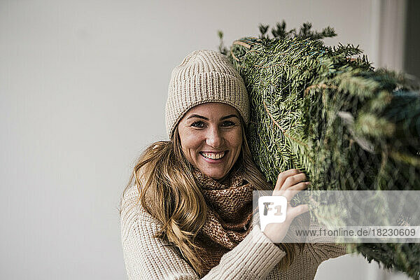 Smiling beautiful blond woman carrying Christmas tree at home