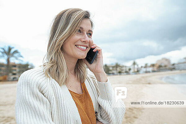 Lächelnde schöne junge blonde Frau  die am Strand mit ihrem Smartphone spricht
