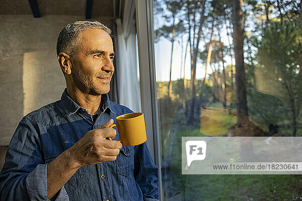 Reifer Mann hält Kaffeetasse zu Hause und schaut aus dem Fenster
