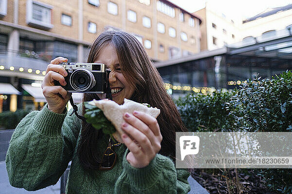 Happy woman holding sandwich photographing through camera in front of building