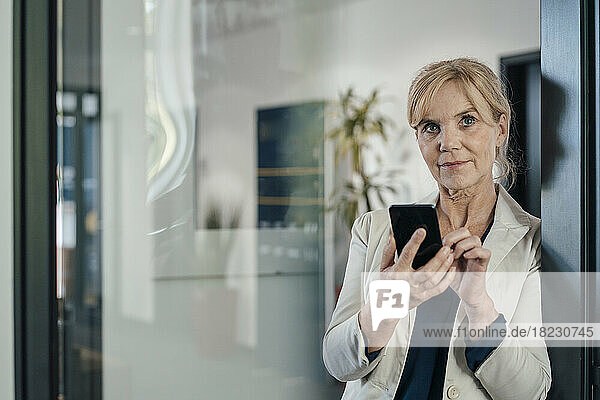 Mature businesswoman with mobile phone in office seen through glass