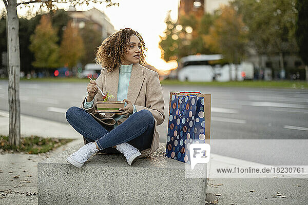 Woman having food sitting with shopping bags on concrete at footpath