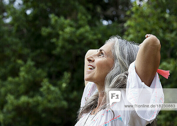 Happy mature woman with hands behind head at forest