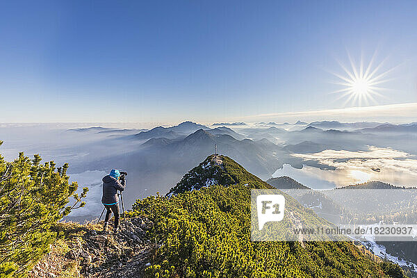Germany  Bavaria  Woman photographing surrounding landscape from summit of Herzogstand mountain at sunrise