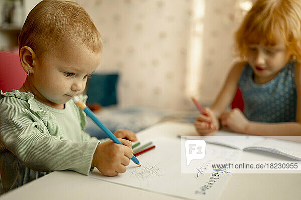 Cute baby girl drawing on paper with sister doing homework at table