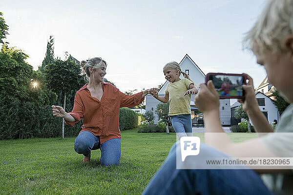 Boy photographing mother dancing with girl through mobile phone in garden