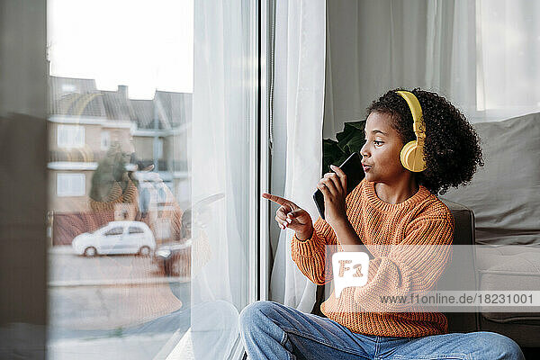 Girl wearing wireless headphones sitting with smart phone and gesturing at window