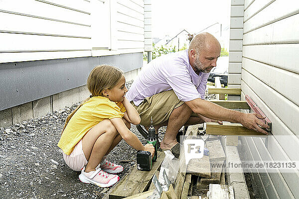 Father using leveling tool crouching with daughter near house